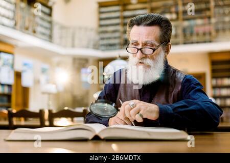 Old man with gray beard, in glasses, vintage clothes, reading a book in the ancient library, using magnifying glass. Close up portrait
