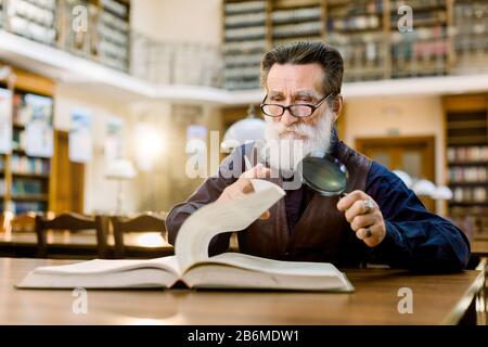 Senior caucasian bearded man in glasses and stylish shirt and leather vest sitting at the table in vintage library, holding magnifying glass and Stock Photo