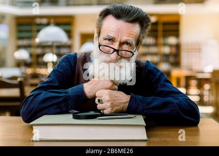 Close up portrait of smiling Caucasian senior bearded man in eyeglasses, sitting at the table with book, magnifying glass and pen, on vintage ancient Stock Photo