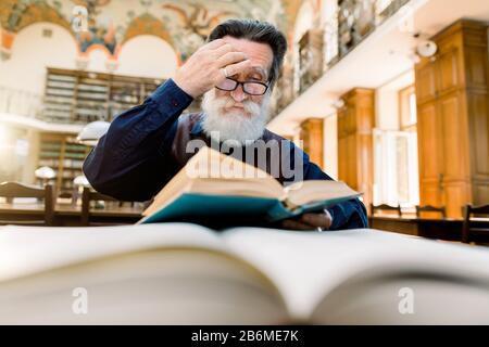 Senior concentrated man with white beard, touching his eyeglasses, sitting at the table and thinking about the book he is reading in the university