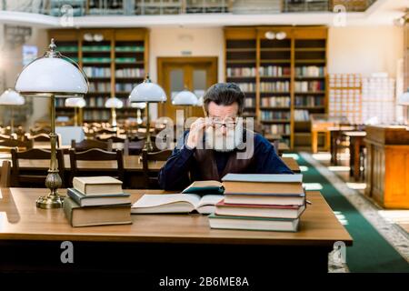 Portrait of elegant senior man, university school teacher, wruter, with beard and eyeglasses, reading books in vintage ancient library on bookshelf Stock Photo