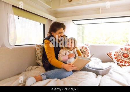 Young woman and little girl smiling and looking at each other while sitting together on cozy sofa next to window with forest view and reading book in Stock Photo