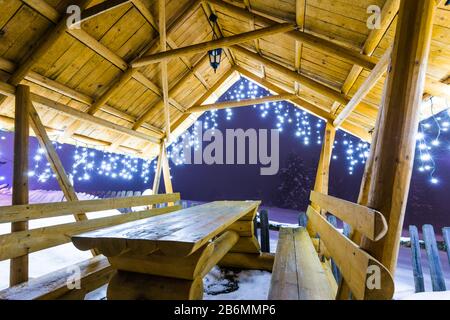 Wooden arbor with Christmas lights stands on a ski slope in the evening foggy winter time against a background of snowy fir trees. Concept of relaxati Stock Photo