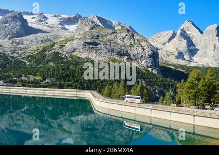 Fedaia Lake, Italy - August 27, 2019: Bus on the Fedaia lake dam, reflected in the turquoise waters, on a bright sunny day, with Marmolada glacier in Stock Photo