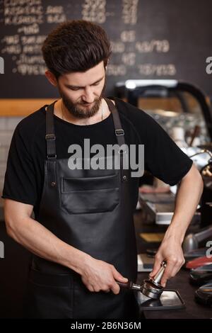 Young male barista pressing ground coffee into bottomless portafilter holder using tamper. Preparing espresso in professional coffee machine. Brewing Stock Photo