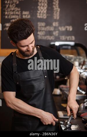 Young male barista pressing ground coffee into bottomless portafilter holder using tamper. Preparing espresso in professional coffee machine. Brewing Stock Photo