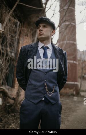 A man posing in the image of an English retro gangster of the 1920s dressed in Peaky blinders style against background of old abandoned building. Stock Photo