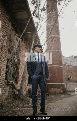 A man posing in the image of an English retro gangster of the 1920s dressed in Peaky blinders style against background of old abandoned building. Stock Photo