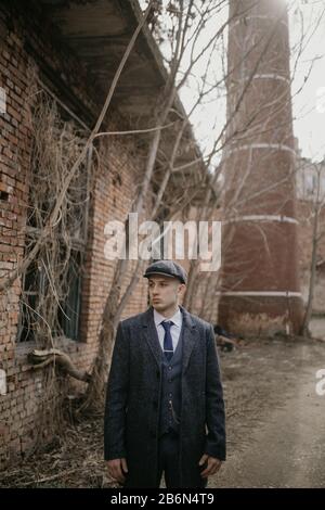 A man posing in the image of an English retro gangster of the 1920s dressed in Peaky blinders style against background of old abandoned building. Stock Photo