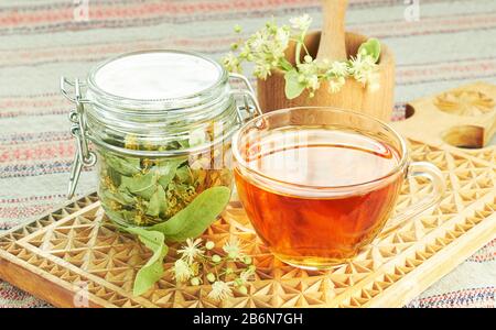Linden tea with lime tree bloom and glass can with dried herb nearby on rustic wooden cutting board, backlit, closeup, copy space, alternative medicin Stock Photo