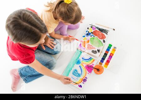 Two children in childrens clothes are sitting on the floor on their knees and enthusiastically painted with bright watercolor paints Stock Photo