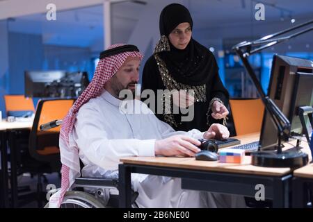 Handicapped Arabic businessman in tradition suit with female arabic assistent working in office. Disabled businessman in the wheelchair works in the o Stock Photo