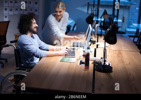 Handicapped young man with female  colleague working in office. Disabled businessman in the wheelchair works in the office at the computer while perfo Stock Photo