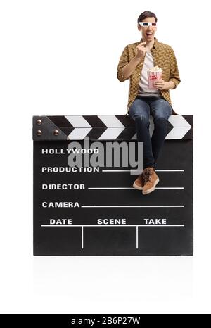 Young man with 3d glasses sitting on a movie clapper board and eating popcorn isolated on white background Stock Photo
