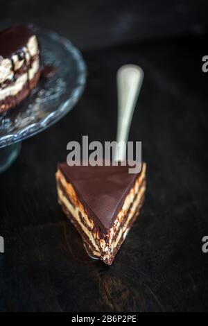 Slice and chocolate cake layers with butter-cream-nut cream with chocolate drips on a glass stand on a dark wooden background. Minimalism, Copy space Stock Photo
