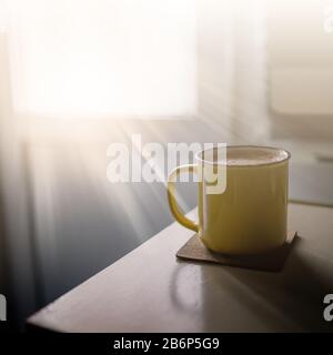 A yellow cup of coffee stands on the office desk in the sunny morning-evening light, next to the monitor and keyboard Stock Photo