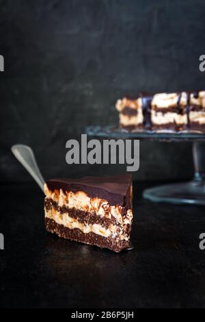 Slice and chocolate cake layers with butter-cream-nut cream with chocolate drips on a glass stand on a dark wooden background. Minimalism, Copy space Stock Photo