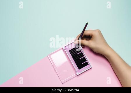 Girl holds tweezers over a pink box with black eyelashes on a pastel background. Woman's hand takes artificial eyelashes for extension. Eyelash extension concept. Stock Photo