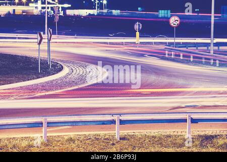 Intersection with car light trails at night, focus on guardrail, color toning applied. Stock Photo