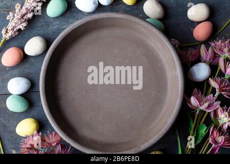 Empty grey Ceramic Plate with painted Easter Eggs of pastel colors on vintage wooden background with various flowers. Happy Easter card concept Stock Photo