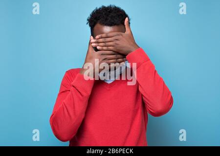 African american black man covering eyes being stressed trying to stay anonym. Studio shot on blue wall. Stock Photo