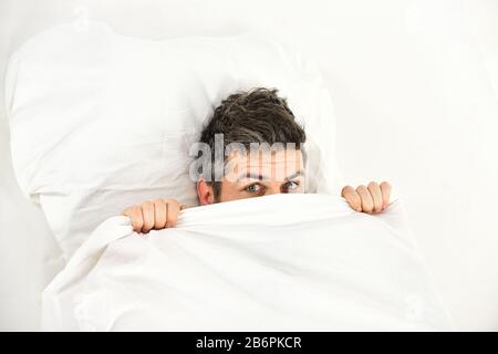 Man with hiding in bed under blanket at home. Guy with surprised or scared face lay under white blanket. Stock Photo