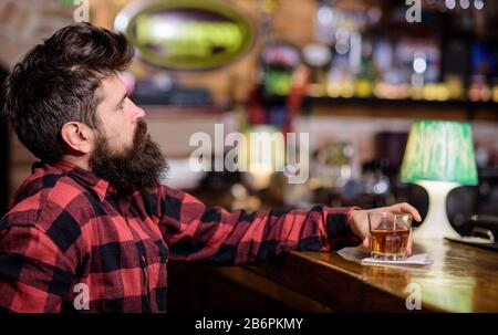 Guy spend leisure in bar, defocused background. Man with dreamy face sits near bar counter. Hipster with beard ordered alcohol drink. Relaxation concept. Man drinks whiskey or cognac Stock Photo