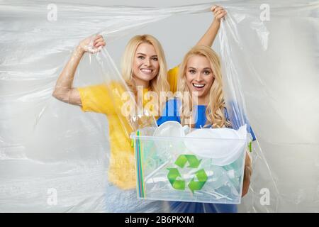 Young women volunteers in yellow and blue t-shirts behing a wall of non-degradable polyethylene wrap trying to fight cellophane. with trash bin. Waste Stock Photo