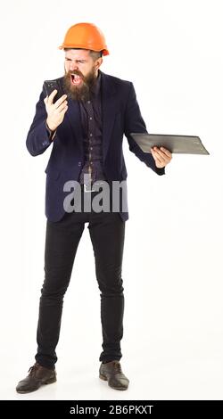 Brutal man wears helmet. Inspector holds documents and talks on phone. Supervisor with papers, helmet and angry face shouting. Angry inspector, management, new technologies, communication concept. Stock Photo