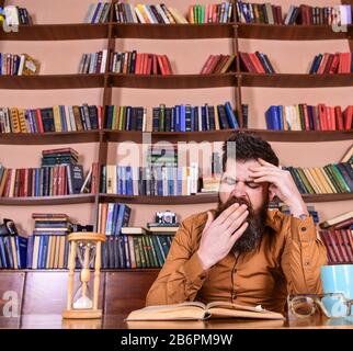 Man on yawning face reading book, studying, bookshelves on background. Man sits at table with mug and hourglass. Teacher or student with beard studying in library. Self education concept Stock Photo