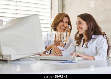 Smiling mid-adult business woman talking on the phone while her young adult colleague types on a computer in an office. Stock Photo
