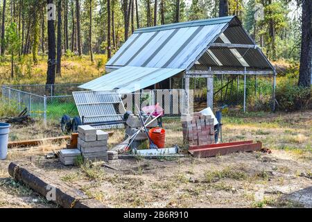 Tools for garden and construction materials sit on a dirt lot with a greenhouse behind in a rural setting. Stock Photo