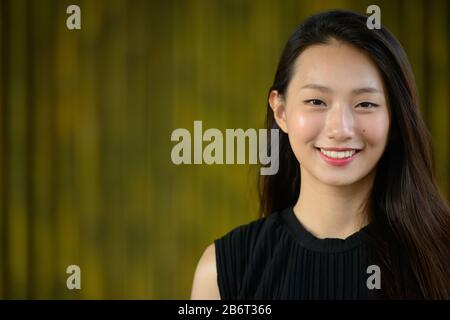 Happy young beautiful Asian businesswoman against bamboo fence Stock Photo