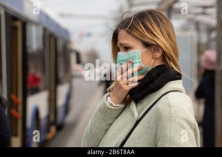 Sad Caucasian woman wearing sterile protective medical mask ill by the Covid-2019 Corona virus at public bus station in a European city street feeling Stock Photo