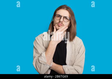 Thoughtful caucasian man with long hair and beard is looking through eyeglasses and touching his chin while posing on a blue wall Stock Photo