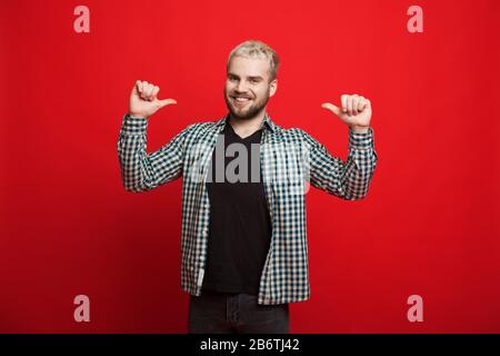 Confident caucasian man with blonde hair and beard is pointing to him while smiling on a red background Stock Photo