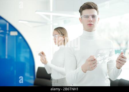 Portrait of serious handsome young man in modern goggles using futuristic tablet while analyzing online scheme Stock Photo