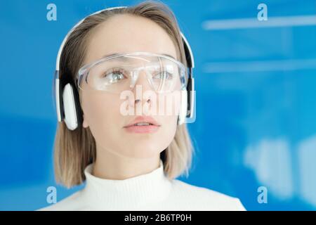 Close-up of pensive young woman in innovative goggles and wireless headphones looking around Stock Photo