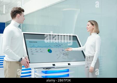 Positive friendly young woman pointing at interactive display and analyzing statistics of product distribution with colleague Stock Photo