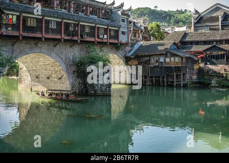Feng Huang, China -  August 2019 : Group of tourists sailing in old historic wooden boat under the landmark arched bridge on the Tuo river, flowing th Stock Photo