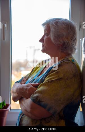 A gray-haired elderly woman stands at the window with her arms crossed over her chest.  Stock Photo