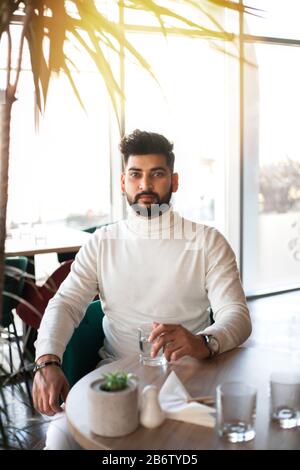 Young indian man drinking water in cafe and looking at camera. Stock Photo