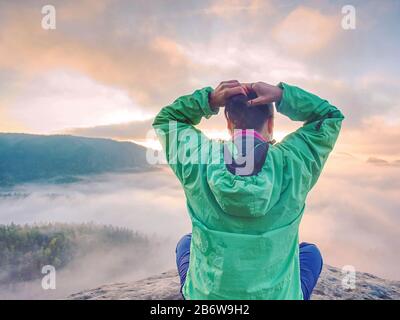 Young woman sit down on top of a mountain celebrate the day. Young lady hiker sitting in blue green windbreaker with red rucksack on sharp cliff Stock Photo