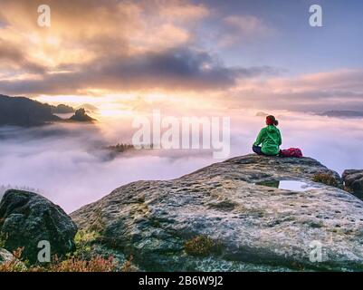 rear view of hiker woman on top of mountain peak in rocks sitting in silence. Thick fog covered the rest of the world Stock Photo