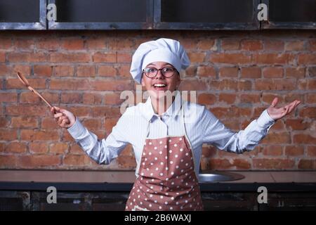 Smiling teenage girl in a cook suit with an apron cap, holds a spoon and shrugs. From the kitchen in the background. Stock Photo