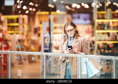 Serious young woman in eyeglasses standing at railing and checking smartphone in mall Stock Photo