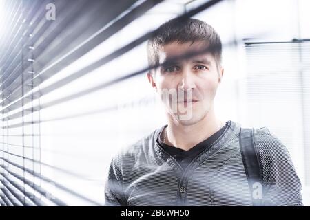 serious young man looking through window blind Stock Photo