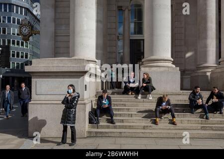 On the day that Chancellor of the Exchequer Rishi Sunak unveiled a £30bn package to boost the economy and get the country through the coronavirus outbreak, a lady wearing a surgical mask makes a call alongside others enjoying early Spring sunshine on the steps of Royal Exchange in the capital's financial district, as the Bank of England's governor Mark Carney cut the interest rate from 0.75% to 0.25%, on 11th March 2020, in the City of London, England. Stock Photo