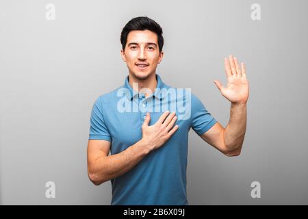 I swear. handsome young man in blue shirt with hand on chest giving oath against grey background. studio shoot Stock Photo