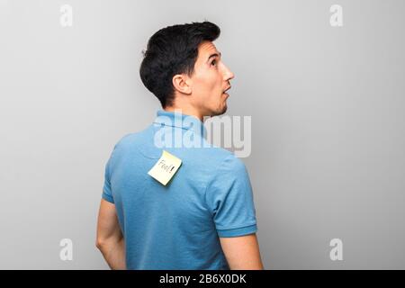 stupid looking man in blue shirt with a yellow sticky note with word fool on his back stupidly looking up Stock Photo